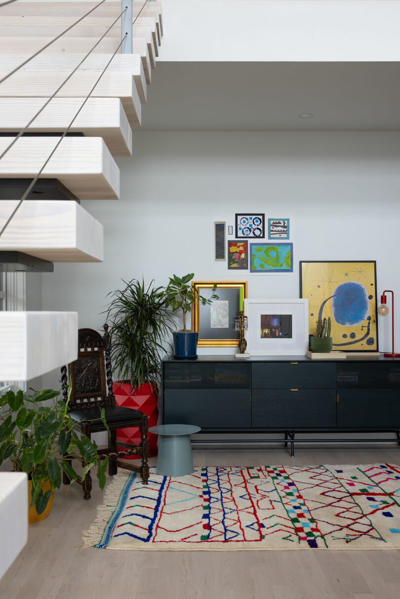 A modern living room with a wall-mounted art collection, plants, a colorful rug, and a staircase. A dark sideboard sits beneath the artwork.