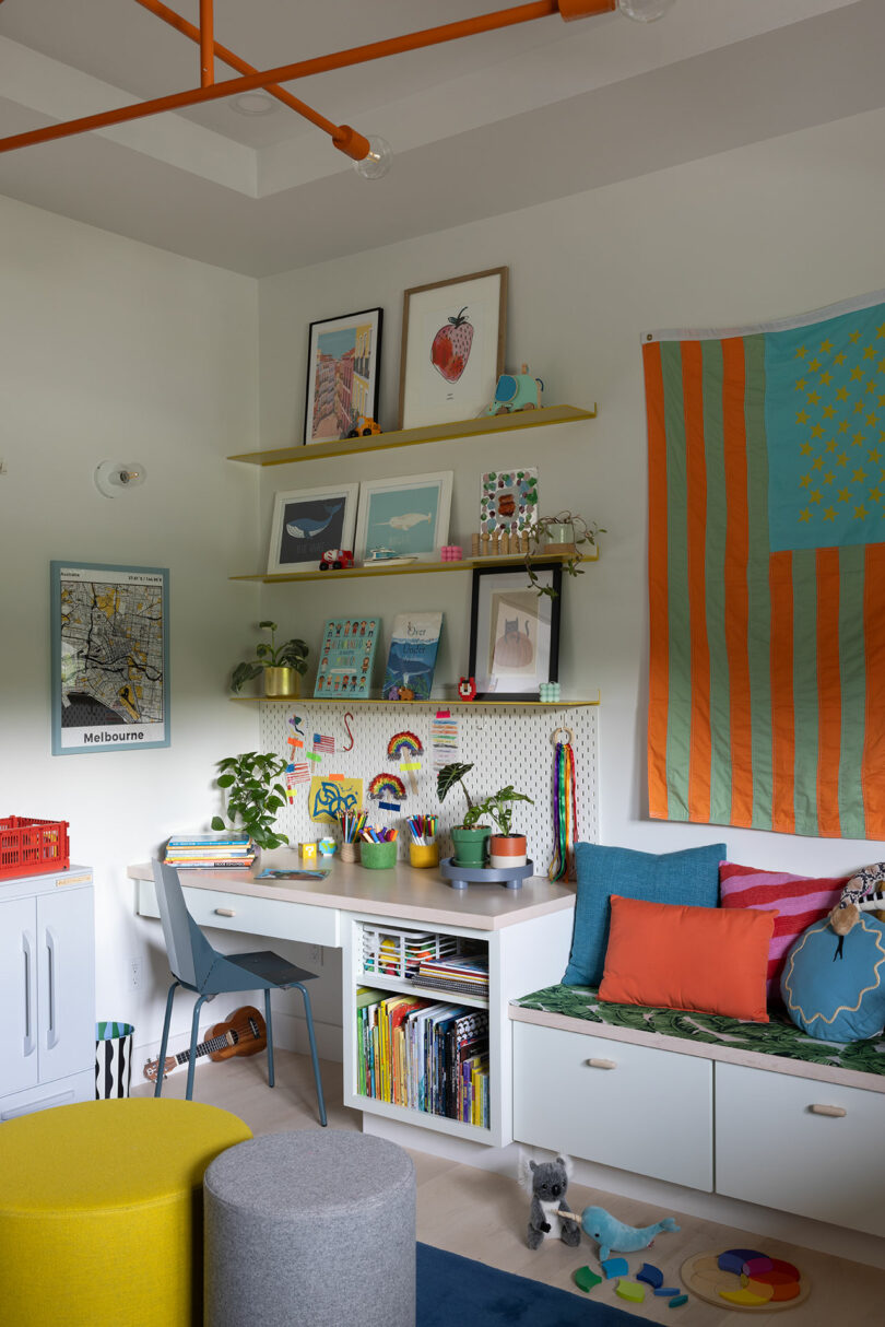 Colorful children's room with a desk, shelves displaying art, a flag tapestry on the wall, and a cushioned bench with toys and books.