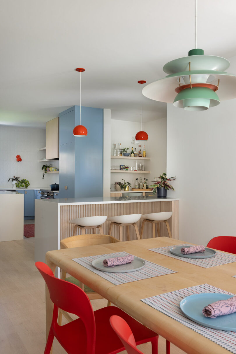 A modern kitchen with colorful pendant lights, a blue accent wall, bar stools at a counter, and a dining table with red chairs and patterned napkins.