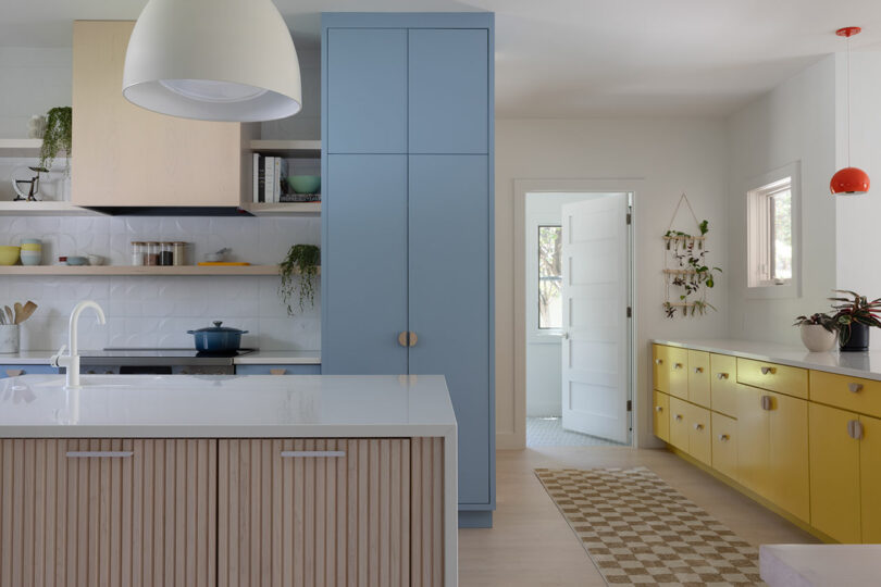 Modern kitchen with a white island, blue cabinet, and yellow lower cabinets. A checkered rug leads to an open door, with plants and shelves adding decor.