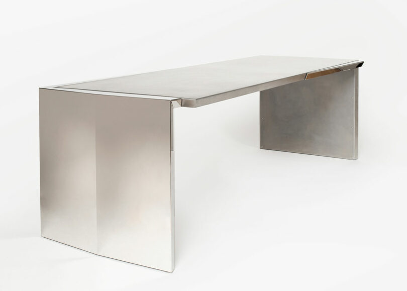 Sleek metallic modern desk with angled sides and a smooth surface, set against a plain background.