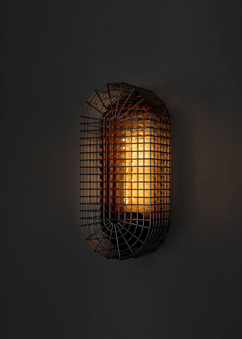 A wall-mounted light fixture with a grid-like metal cage and a warm glowing bulb inside against a dark background.