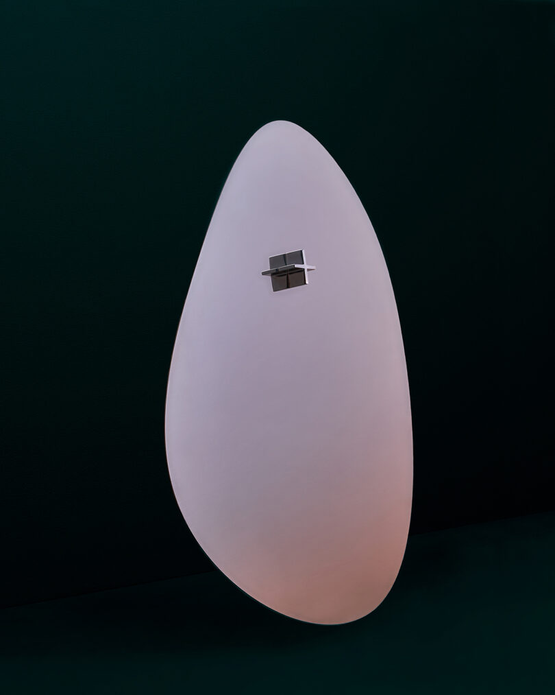 A modern white oval-shaped mirror with a small square metal attachment against a dark green background.