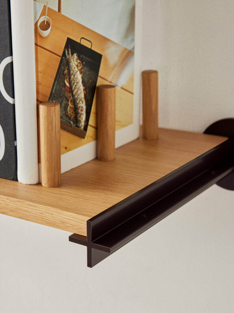 A close-up of a wooden shelf with three cylindrical wooden bookends holding up several cookbooks, one of which has an image of a cooked fish on the cover. A dark brown metal support is visible.