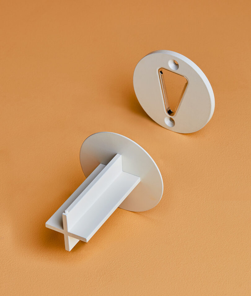 Two parts of a white, circular wall bracket are shown on an orange surface. One piece is flat with two screw holes and the other has a protruding rectangular support.