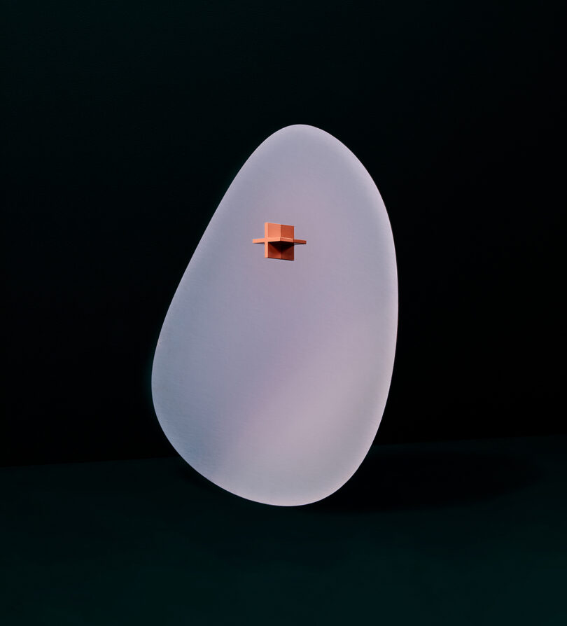 A modern mirror with a smooth, asymmetrical oval shape in pale pink, featuring a small, protruding orange cube in the center, set against a dark background.