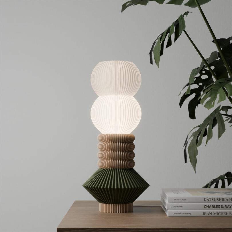 The STAK Modular Lamp, featuring a geometric wooden base and a white spherical shade, elegantly complements the wooden table. It's perfectly placed beside a stack of books and a vibrant green plant.