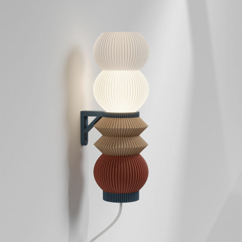 Introducing the STAK Modular Lamp—a wall-mounted fixture featuring three stacked, pleated shades in white, beige, and red, all elegantly supported by a dark metal bracket.