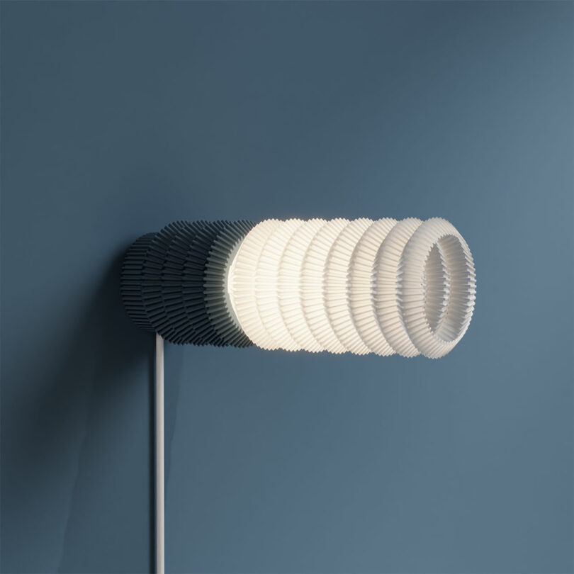 The STAK Modular Lamp features a modern, wall-mounted light fixture with a layered, cylindrical design set against a blue background.