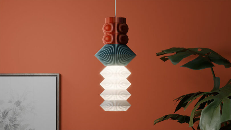 A STAK Modular Lamp, featuring geometric red, blue, and white sections, hangs elegantly in front of an orange wall. A plant and framed artwork are partially visible in the background.