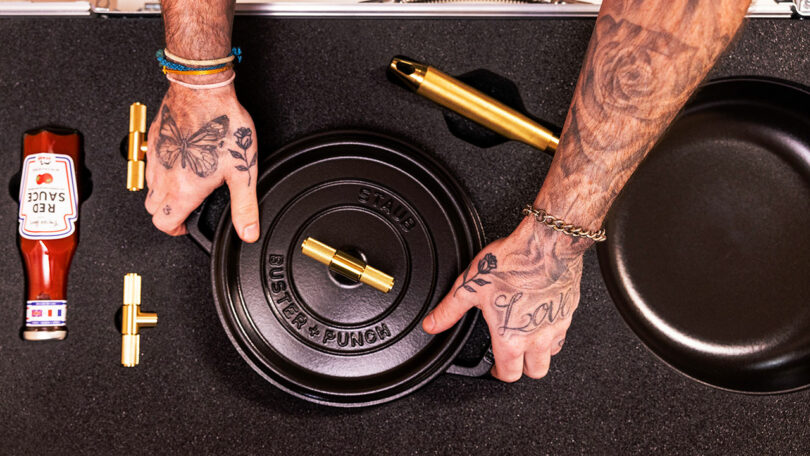 Two tattooed hands handle a cast iron pan lid with the text "Buster + Punch." A black Staub frying pan and bottle of red sauce are nearby on a black surface.