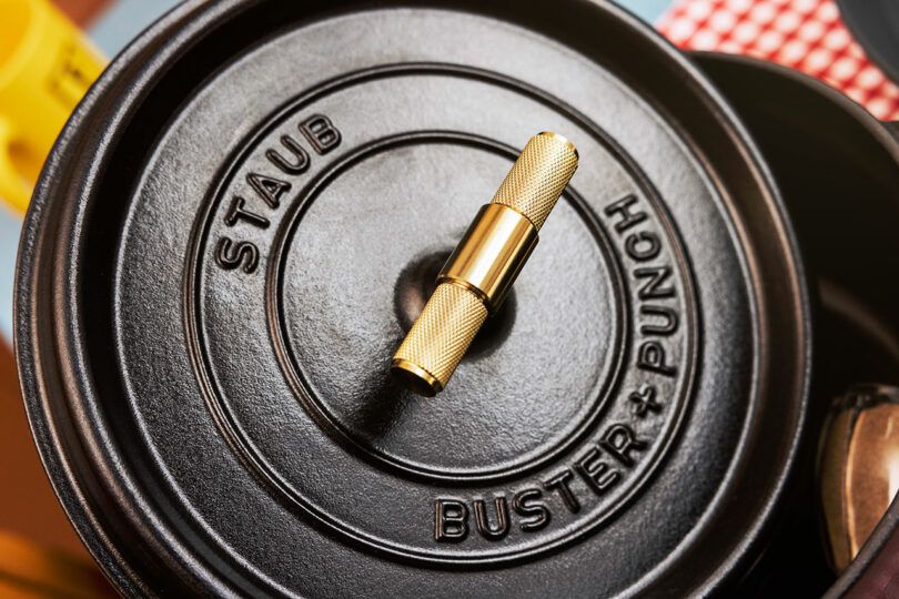 Close-up of a black Staub pot lid featuring a gold handle and the embossed Buster + Punch logo.