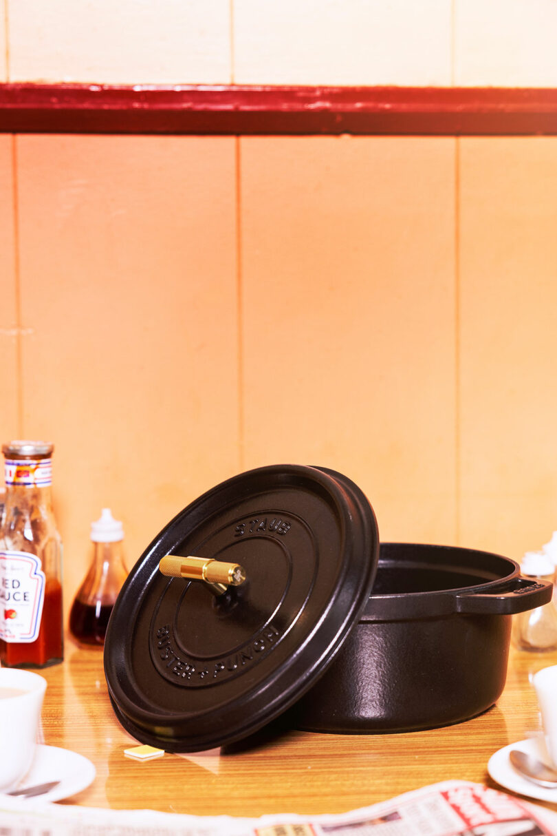 A Staub black cast iron pot with a lid sits on a wooden table, accompanied by condiment bottles, two cups, and a newspaper.