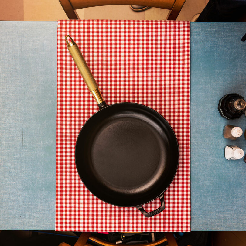 A black frying pan with a golden handle, possibly a Staub, rests on a red and white checkered cloth on a blue table.