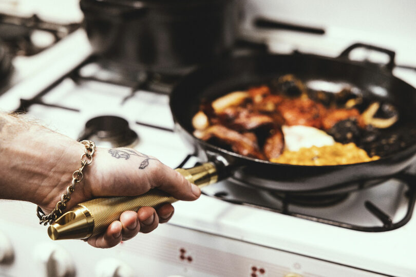 A person cooks food in a black Staub frying pan on a stove, holding the pan by its brass handle.