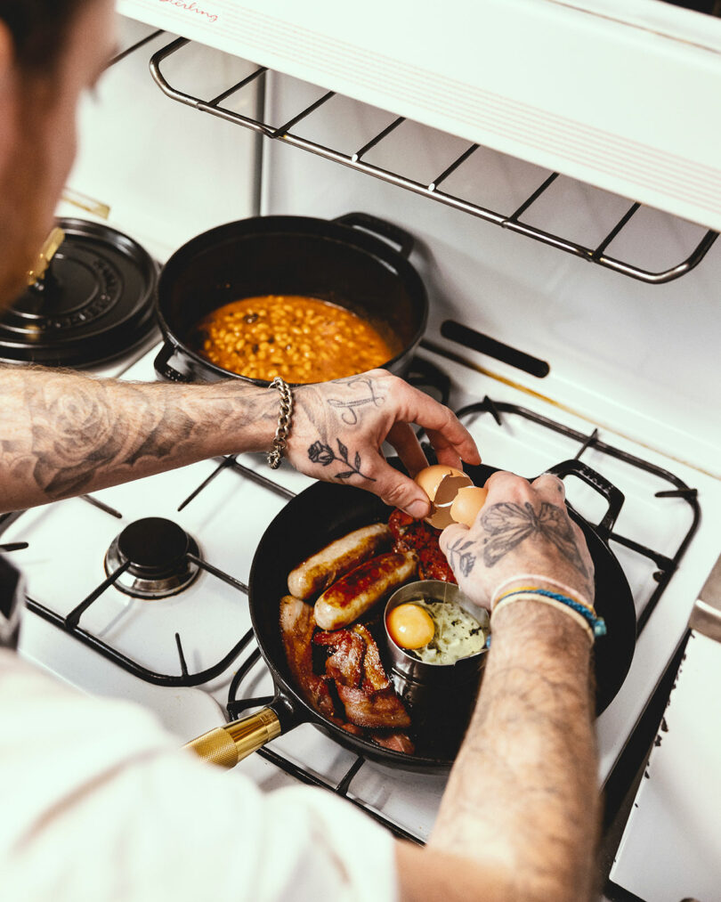 A person with tattoos on their arms is cracking an egg into a skillet on a stovetop, right next to a Staub pot of beans.