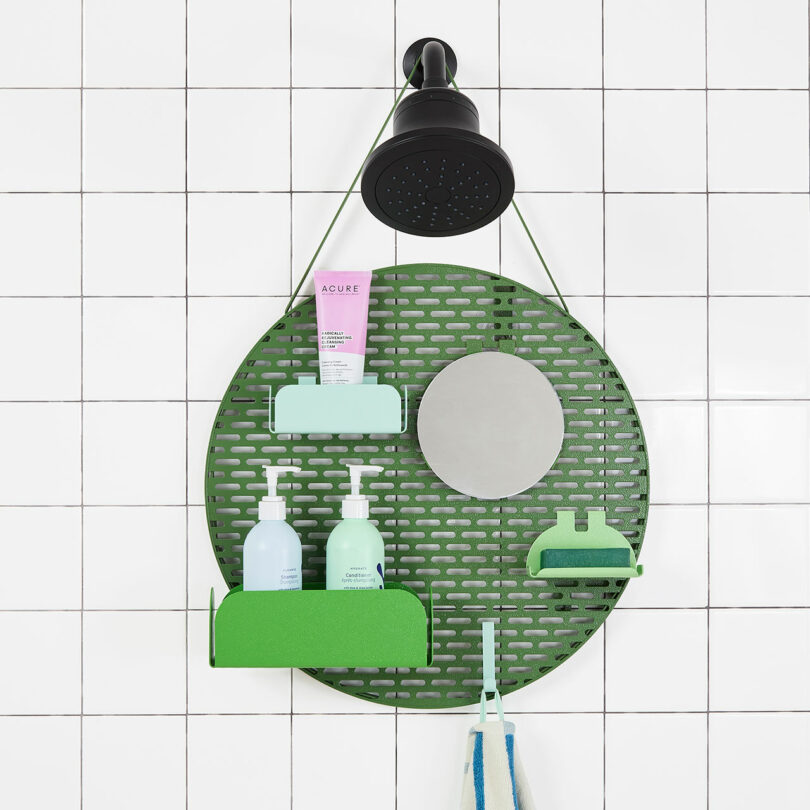 Shower wall with a green circular hanging organizer holding various toiletries, including bottles of soap and shampoo, a tube of cream, and a small mirror. A black showerhead is visible above.