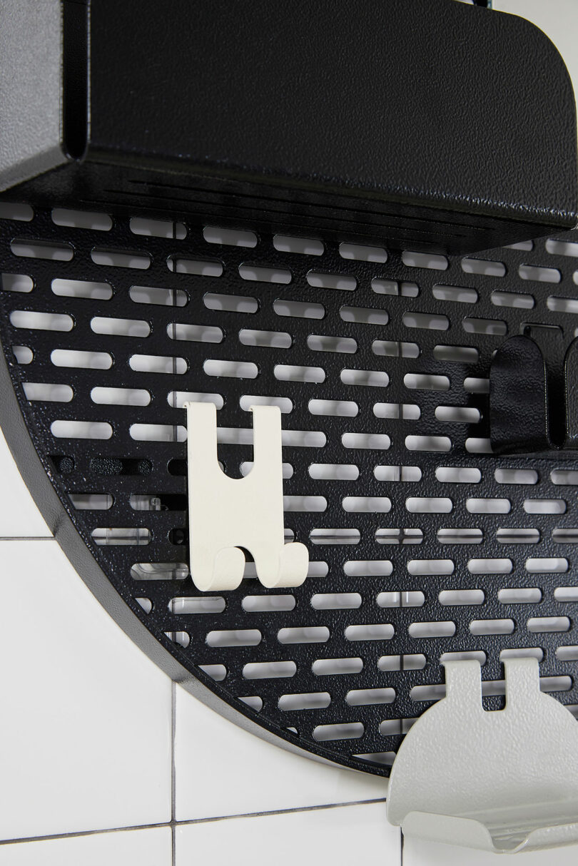 A black perforated wall organizer with two white hooks mounted on a tiled wall.