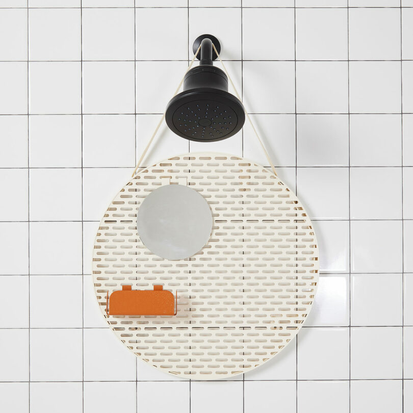 A round shower caddy with small holes is hanging on a tiled wall. It holds a round mirror and an orange bar of soap. A black showerhead is mounted above it.