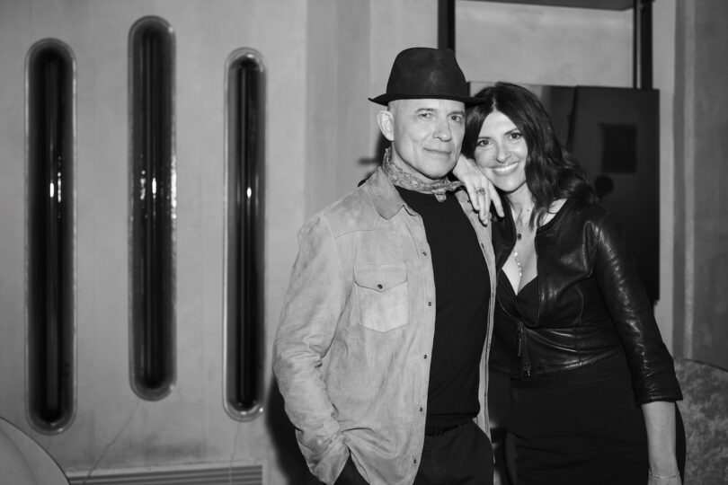 Black and white photo of a man in a hat and a woman in a leather jacket standing together indoors, with the woman smiling and her arm around the man's shoulder