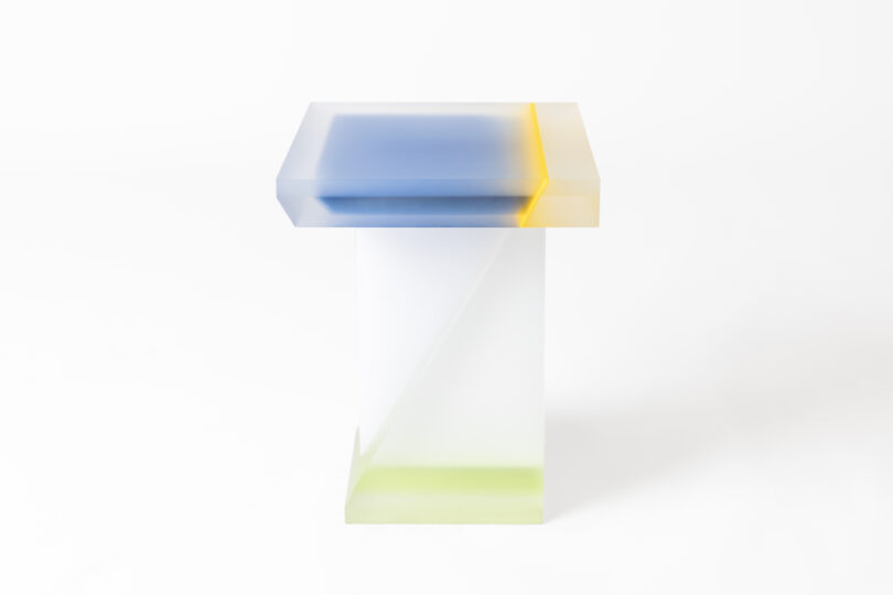 A modern, semi-transparent table with a gradient of blue, yellow, and green colors, featuring a geometric design with a square top