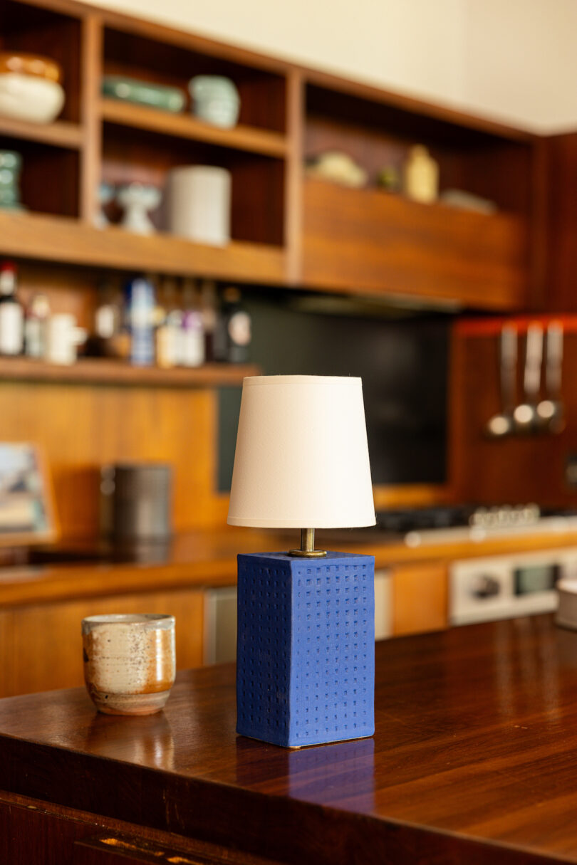 A small blue table lamp with a white shade sits on a wooden countertop in a kitchen. Shelves, kitchen utensils, and other items are visible in the background