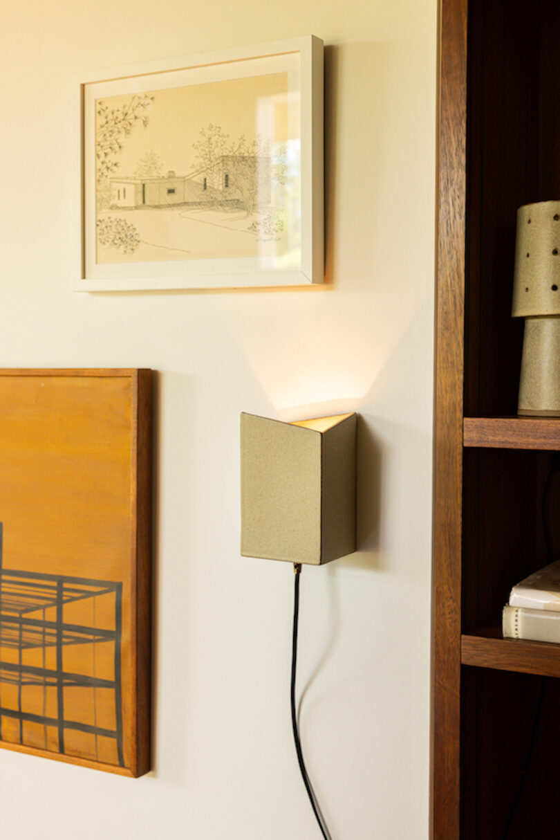 A modern wall sconce emits light beneath a framed architectural drawing and beside a wooden shelf with books