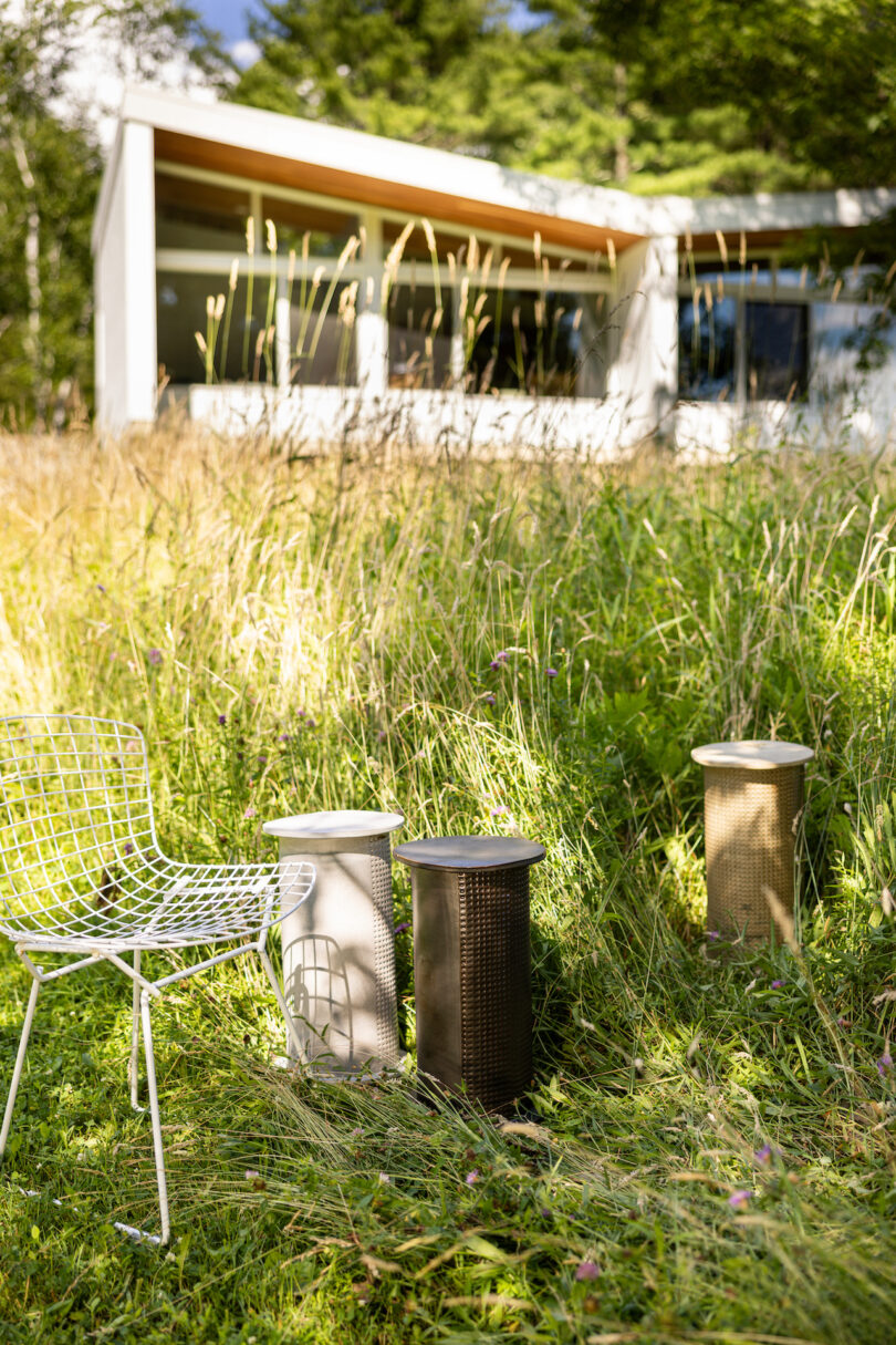 A modern white chair and three cylindrical outdoor heaters are placed on grassy ground with a contemporary house in the background amidst tall grass and trees