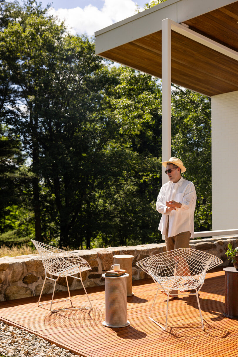 A person wearing a hat and sunglasses stands on an outdoor wooden deck with two wire chairs, a small table, and a lush green background