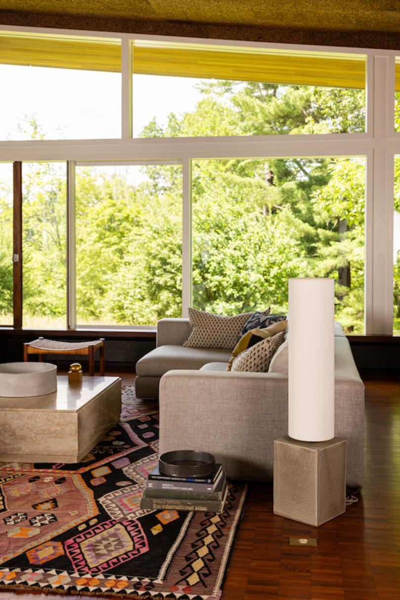 A modern living room with a gray sofa, a colorful patterned rug, a concrete coffee table, and a large floor lamp. Floor-to-ceiling windows offer a clear view of the lush greenery outside