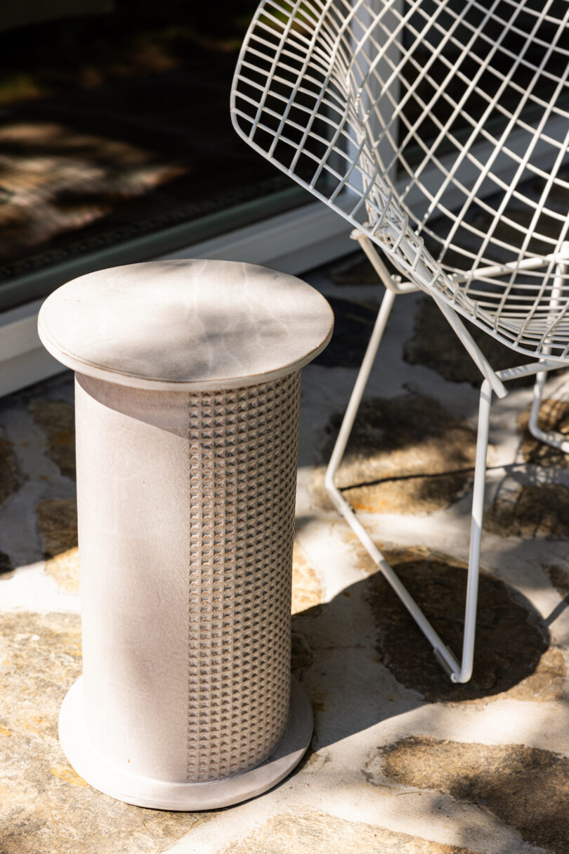 A textured cylindrical side table beside a white wireframe chair is placed on a stone-paved surface, illuminated by sunlight