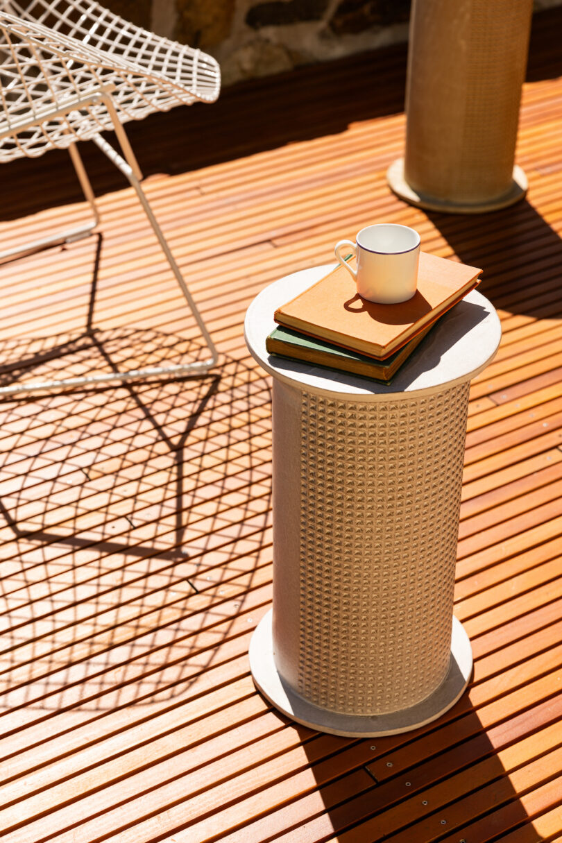 A white metal side table on a sunlit wooden deck holds a stack of books and a white mug. An empty white metal chair casts a shadow nearby