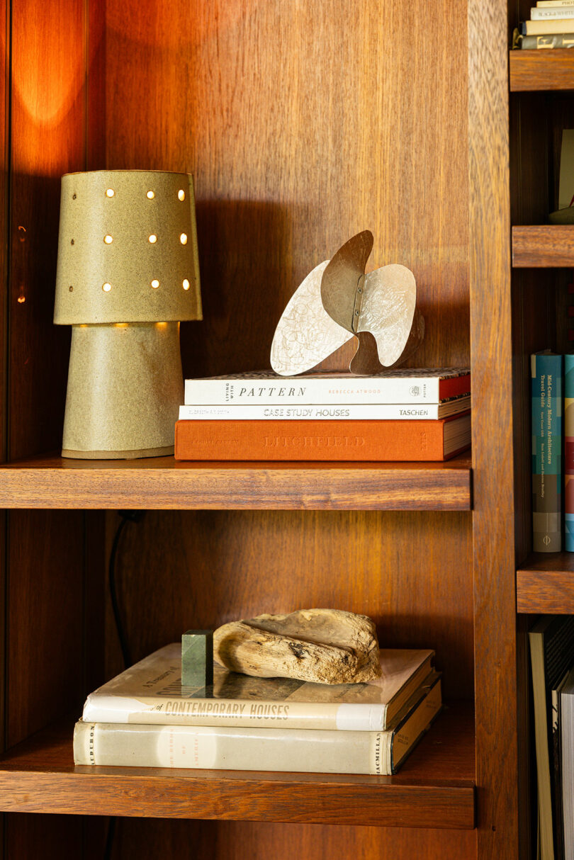 A wooden bookshelf displays a lamp with small holes, a decorative object of abstract shapes, and several stacked books, exuding a touch of Modern Bohemia.