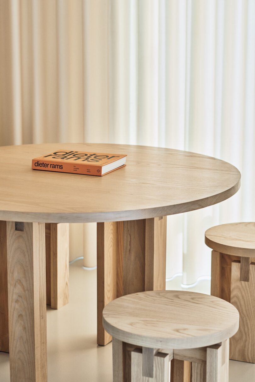 A wooden table with two matching wooden stools placed around it. A book titled "Dieter Rams" rests on the table. A light curtain hangs in the background