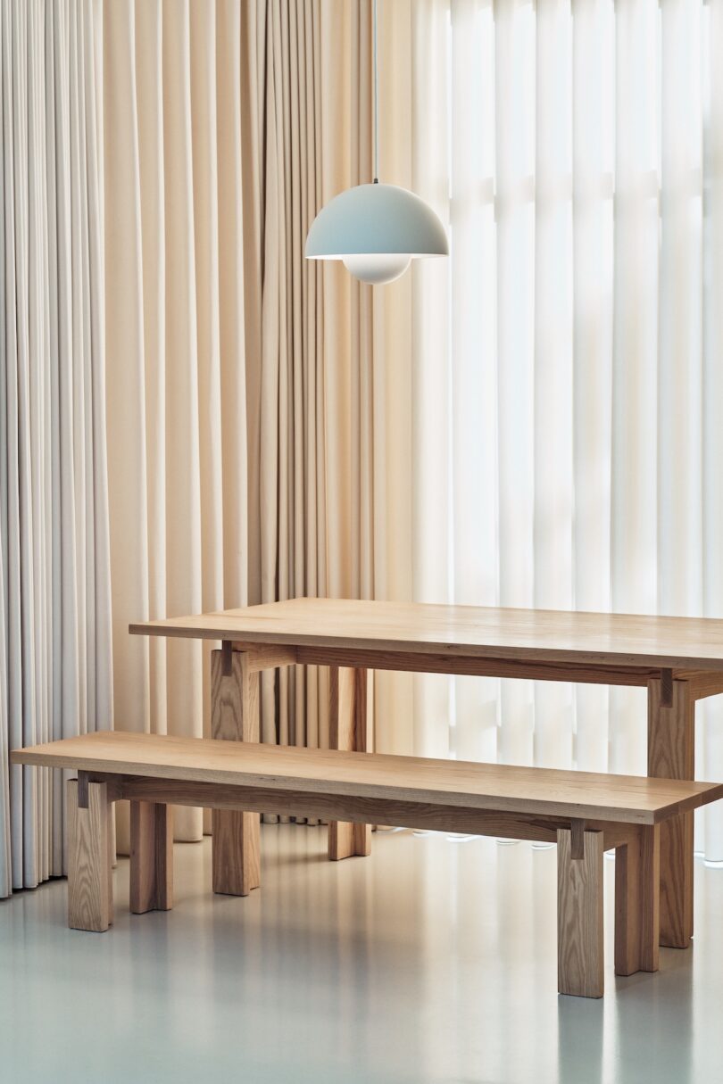 A minimalist dining area with wooden table and bench, surrounded by tall beige curtains, under a white dome-shaped pendant light