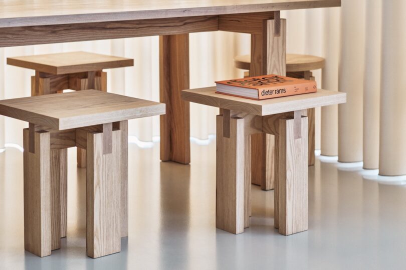 A set of wooden minimalist furniture including a table and small stools. One stool has an orange book titled "Dieter Rams" on it. The background features light-colored curtains