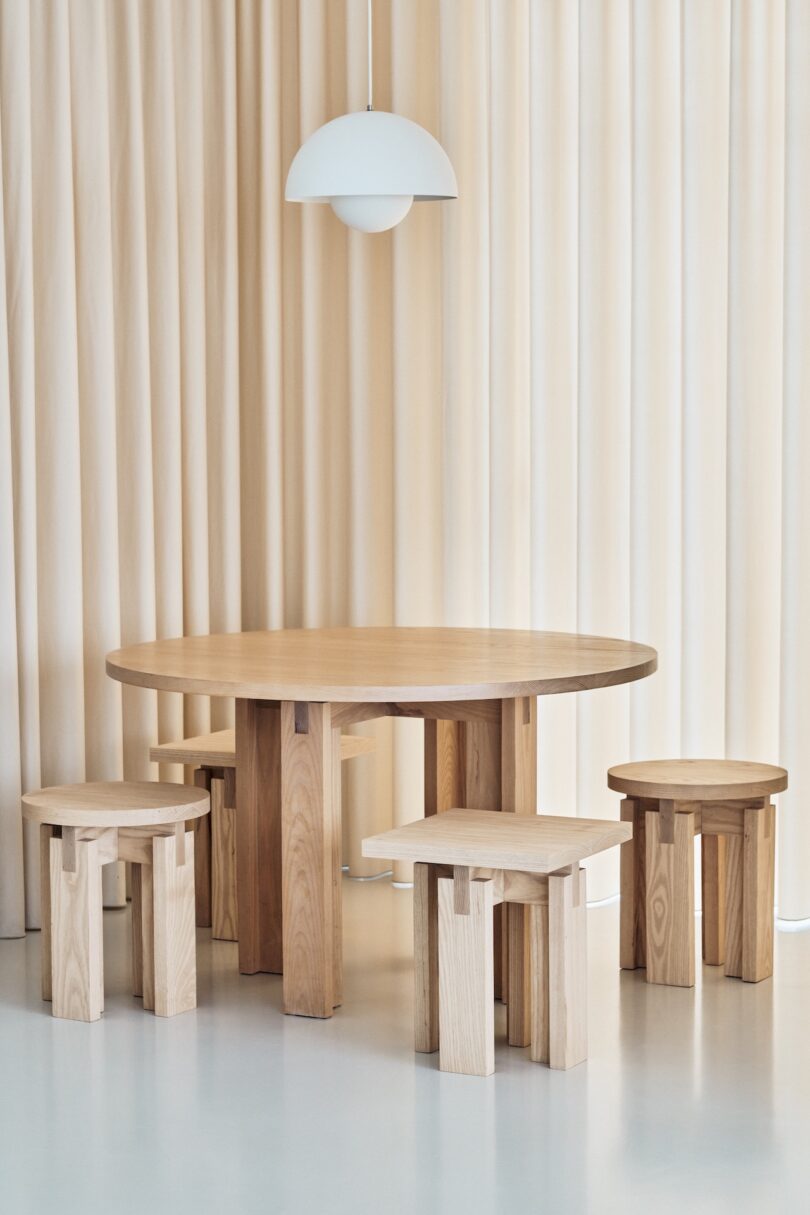 A round wooden table with five matching wooden stools is set against a backdrop of beige curtains, under a white pendant light