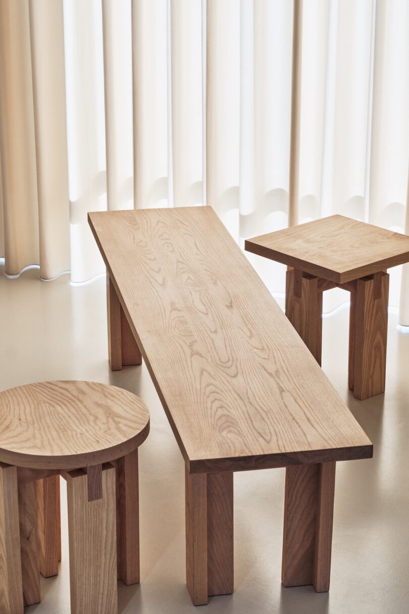 A minimalist room featuring a long wooden bench, a round wooden stool, and a square wooden table, all set against white curtains