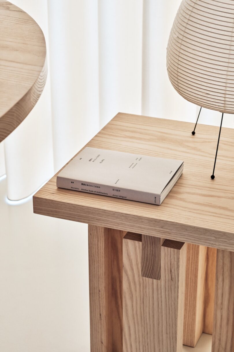 A closed book rests on a light wooden table, next to a spherical lamp with a ribbed paper shade