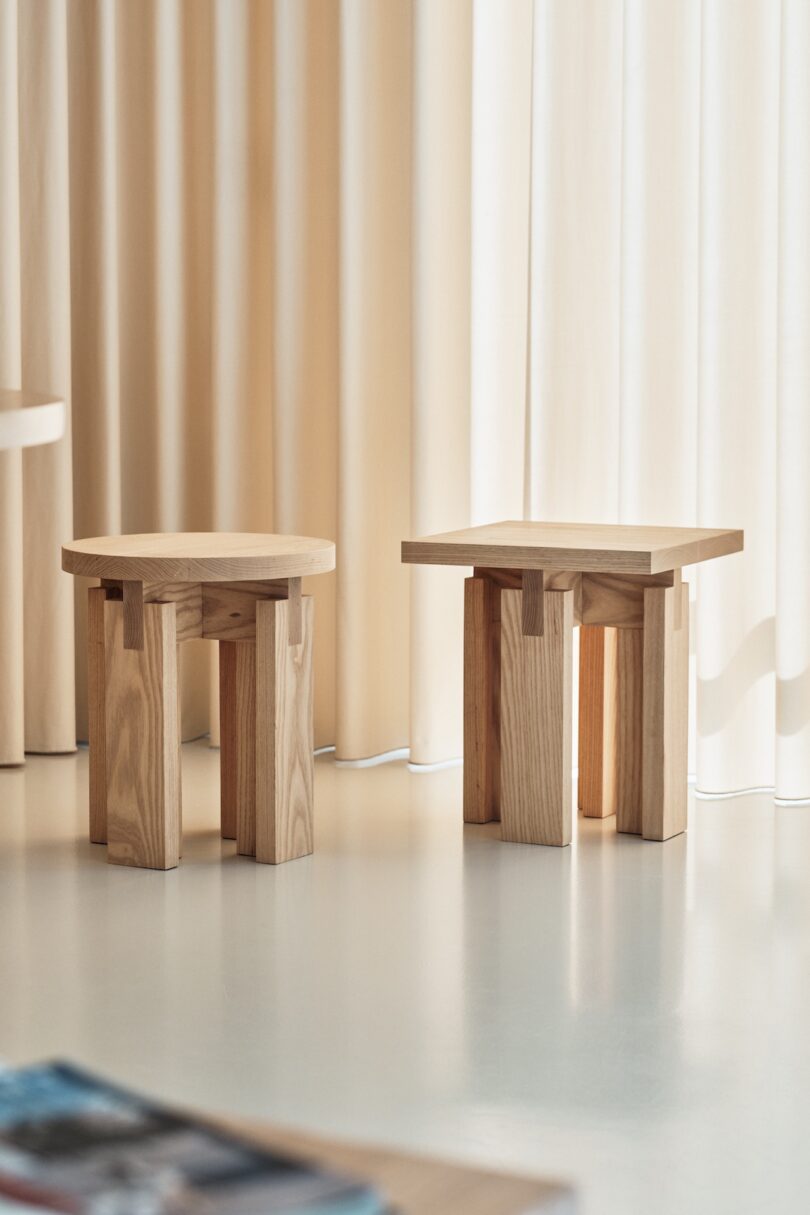 Two wooden stools with unique geometrical designs stand on a light floor near beige curtains