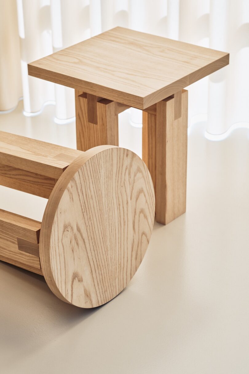 A modern wooden side table and a matching round wooden stool are placed on a light-colored floor with a light-colored backdrop