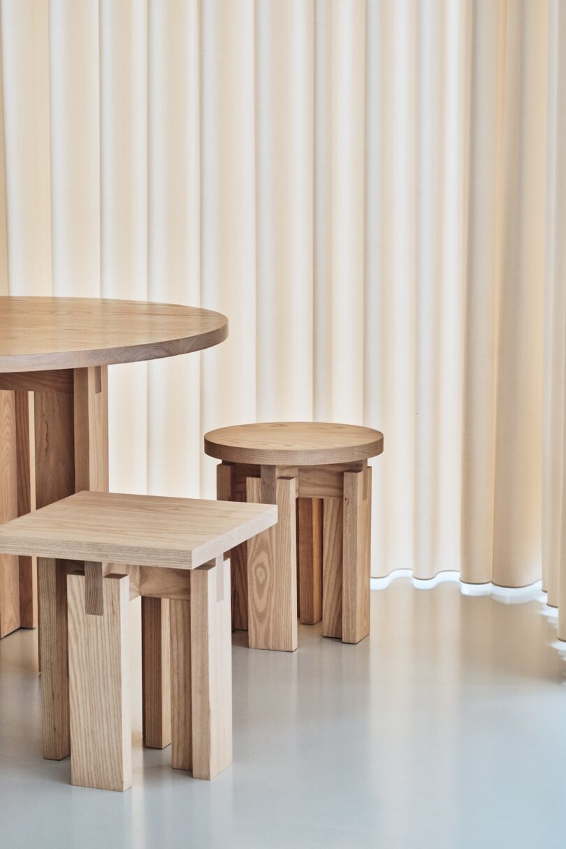 Minimalist wooden furniture set with a round table and two stools on a smooth floor, positioned against a backdrop of beige vertical blinds