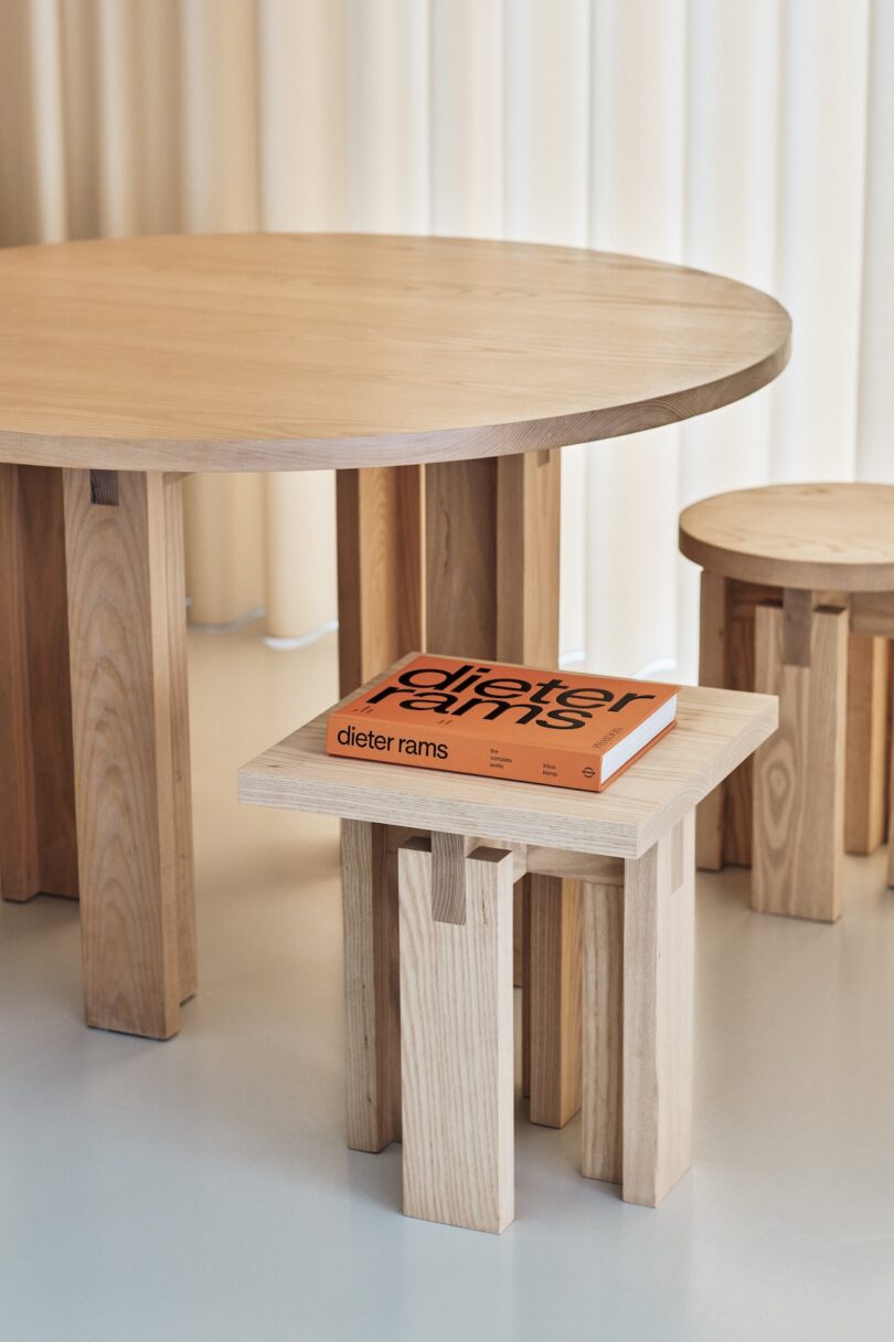 A wooden round table, a small wooden stool, and a rectangular wooden side table with an orange book titled "Dieter Rams" on it are shown in a minimalist setting with a light curtain in the background