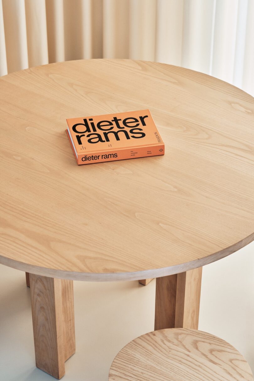 A round wooden table with a book titled "dieter rams" placed on top. A matching wooden stool is partially visible at the bottom