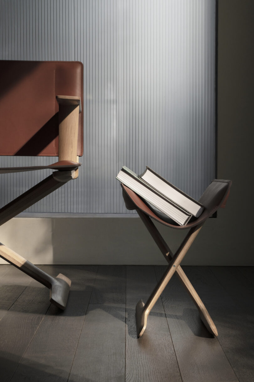 A wooden chair with brown leather backrest and seat stands next to a folded wooden stool holding an open book, set against a ridged glass window under a soft light