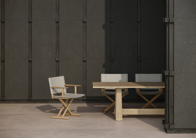 Modern, minimalist room with a wooden table and three chairs, one positioned away from the table. The walls are dark and the floor is a light neutral color