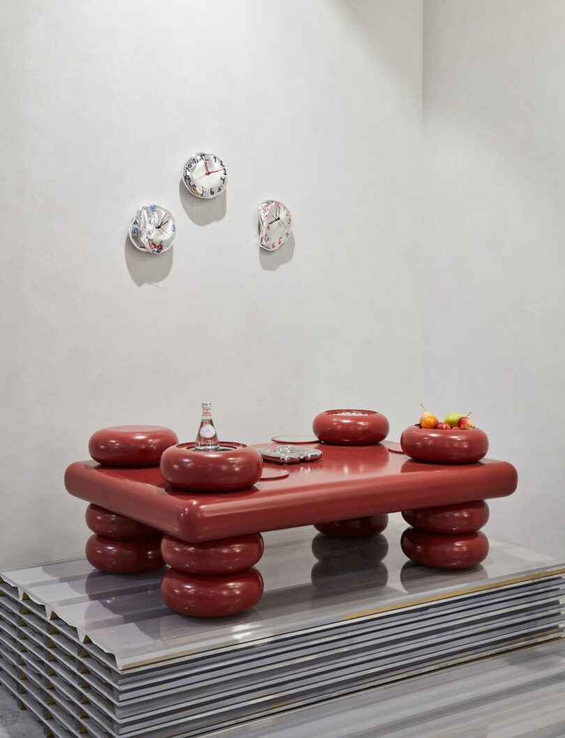 A modern table with a unique design features large, red cylindrical legs and a matching tabletop. It is decorated with a decanter, glass, and fruit bowl. Three abstract wall sculptures hang above it