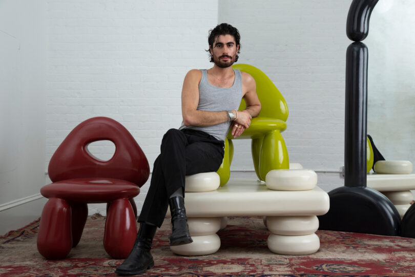 A man is sitting on colorful, modern furniture in a room with white brick walls. He is wearing a gray tank top, black pants, and black boots, with his arms resting casually on the back of a chair