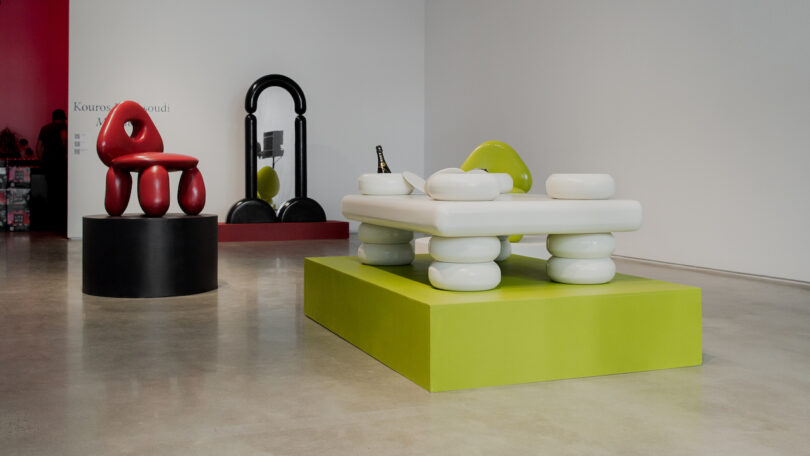 Modern art exhibit featuring abstract furniture: a red chair on a black platform, a green and white chair next to a matching table on a green platform, and a black floor mirror against a red wall