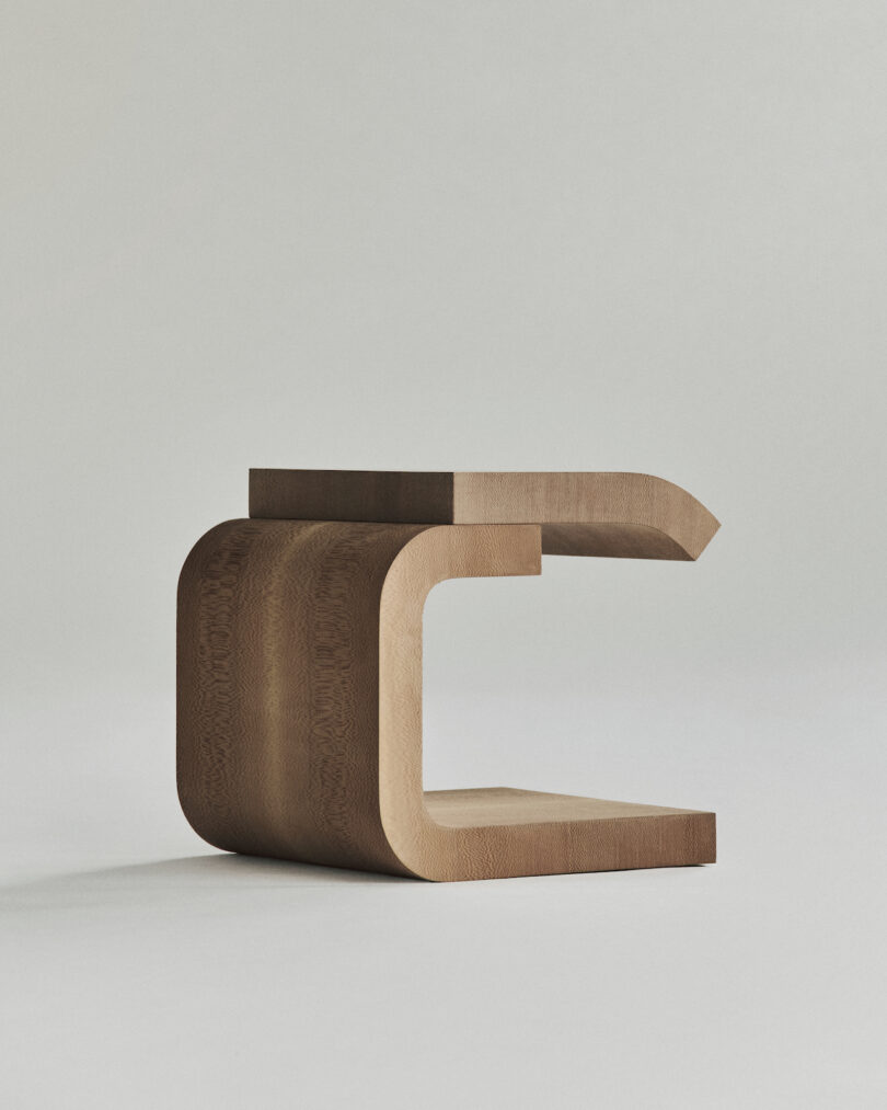 A minimalist wooden table with a C-shaped design stands against a plain background.